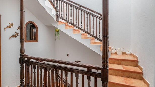 Una escalera con balaustres de madera y barandilla, que conduce a un rellano con pared blanca y una pequeña ventana en arco.