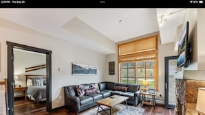 Ein modernes Wohnzimmer mit einer Couch, einem Couchtisch und einem Kamin. Ein Fernseher ist an der Wand montiert und es gibt ein Fenster mit Jalousien.