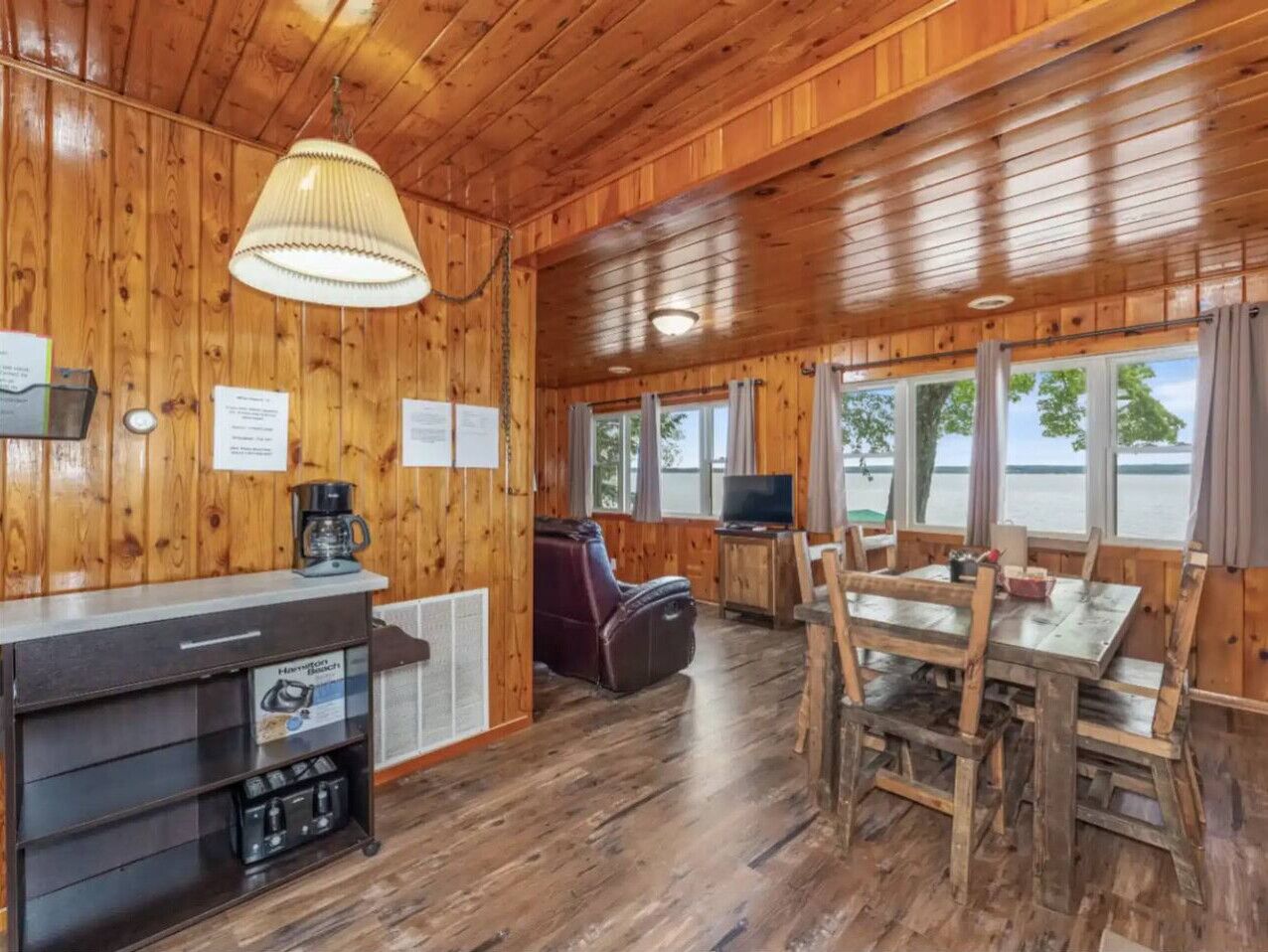 A cabin interior with wooden walls and floors, a dining table with chairs, a sofa, a TV, and a coffee station.