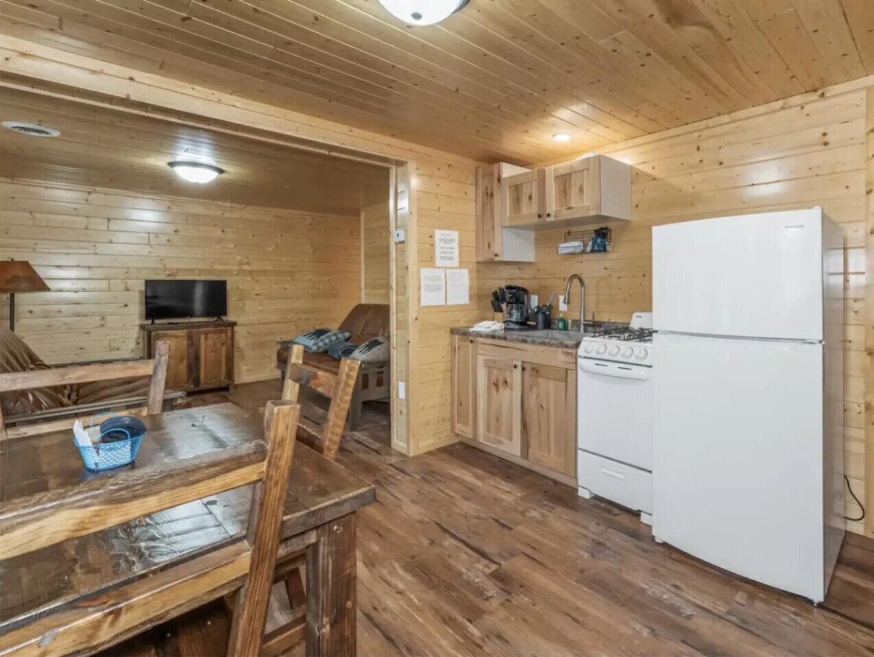 A wooden interior with a dining table, chairs, a kitchen area with a stove and refrigerator, and a television in the corner.