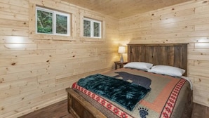 Ein Schlafzimmer einer Holzhütte mit Bett, zwei Fenstern, einem Nachttisch und einer Deckenleuchte.