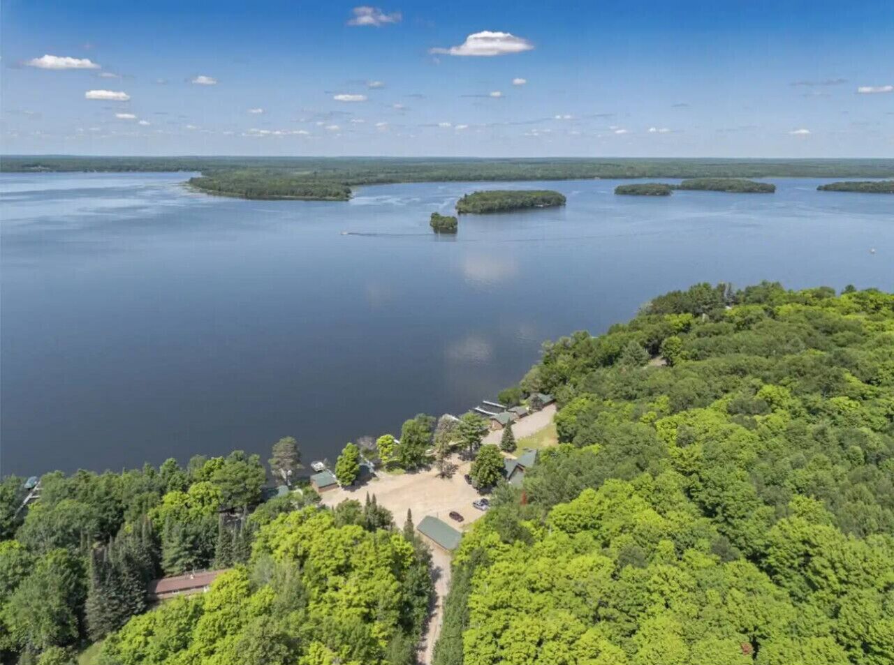 An aerial view of a lakeside property with a parking area, buildings, and dense forest.