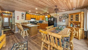 A rustic kitchen with wooden cabinets, a central island, and a dining area with wooden chairs. - Cabin #12 Thunder Bay Resort (Phelps)