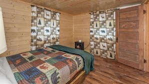 Ein gemütliches Blockhauszimmer mit Holzboden, einem Bett mit bunter Bettdecke, Karo-Vorhängen und einem kleinen Tisch mit Kerze.