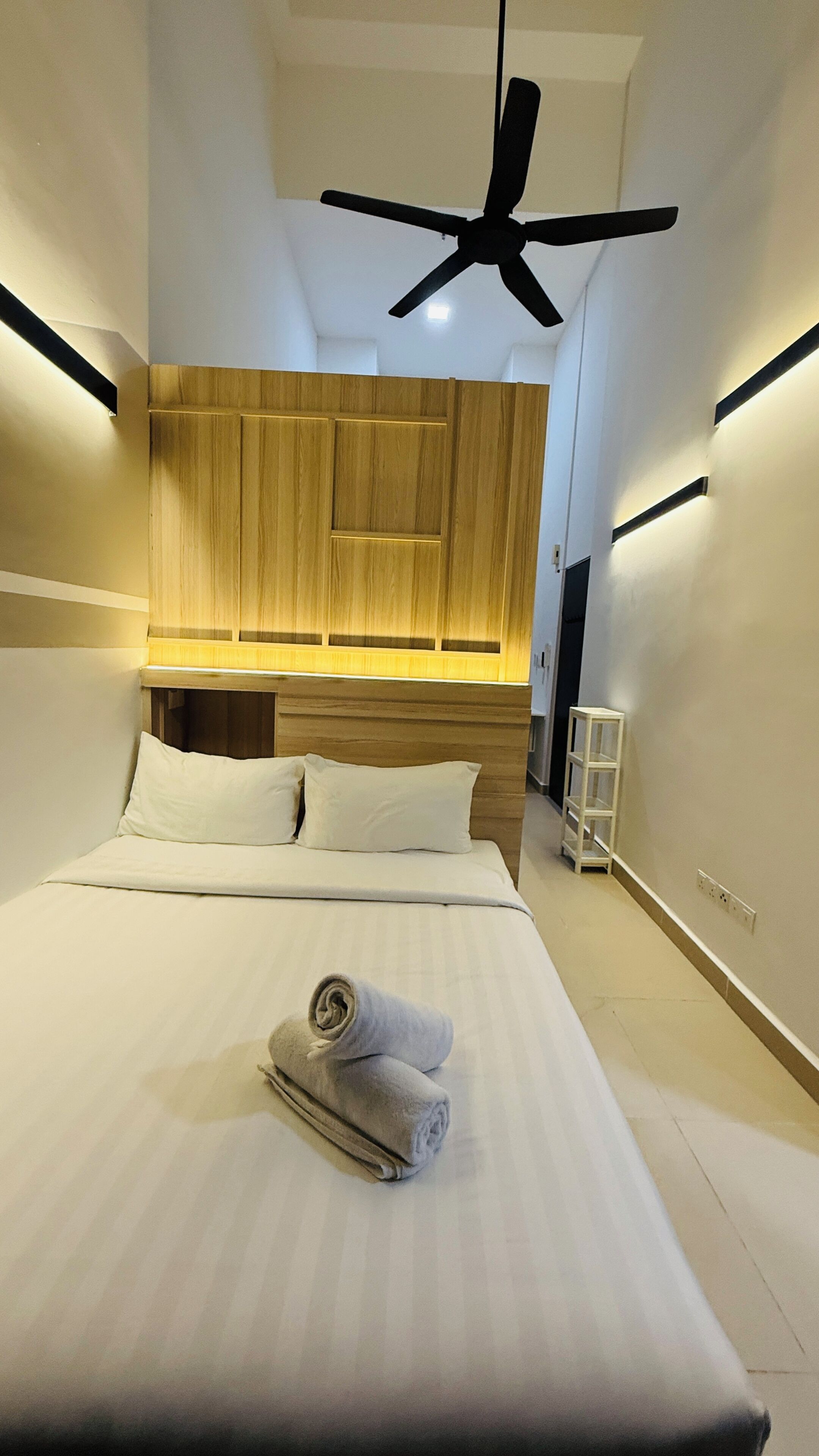Una habitación de hotel moderna con una cama grande, ventilador de techo, cabecero de madera y una mesita de noche blanca.