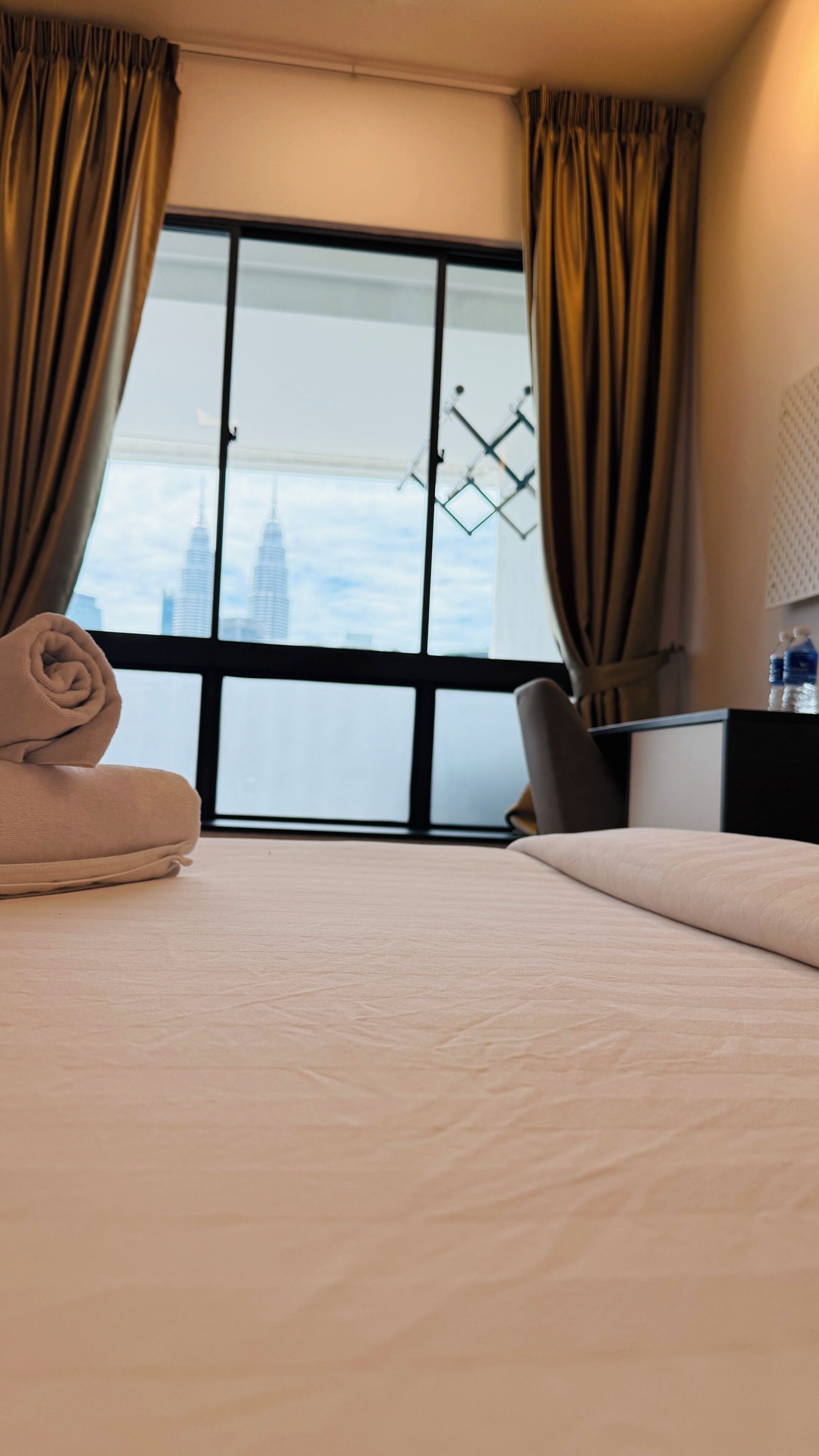 A hotel room with a bed, a desk, a chair, and a view of a cityscape through a large window.