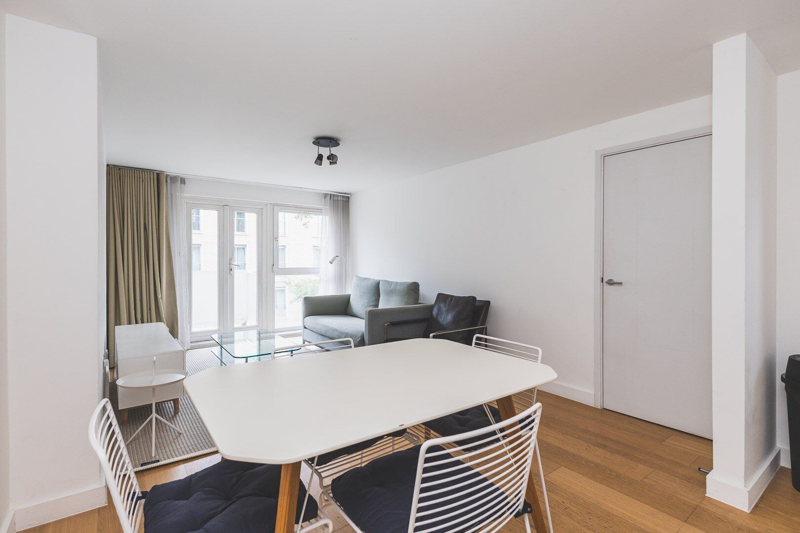Una habitación moderna y luminosa con una mesa blanca, sillas y un sofá. Se ve una puerta y una ventana con cortinas.