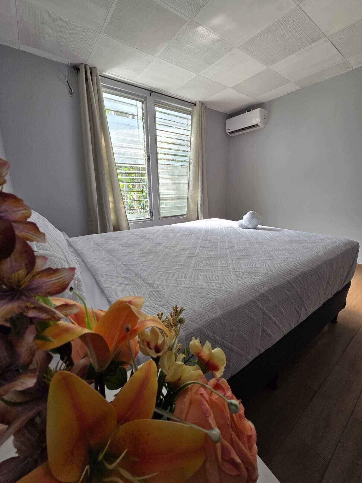 Una habitación de hotel con cama, dos ventanas con persianas, un equipo de aire acondicionado y un florero con flores sobre la mesita de noche.