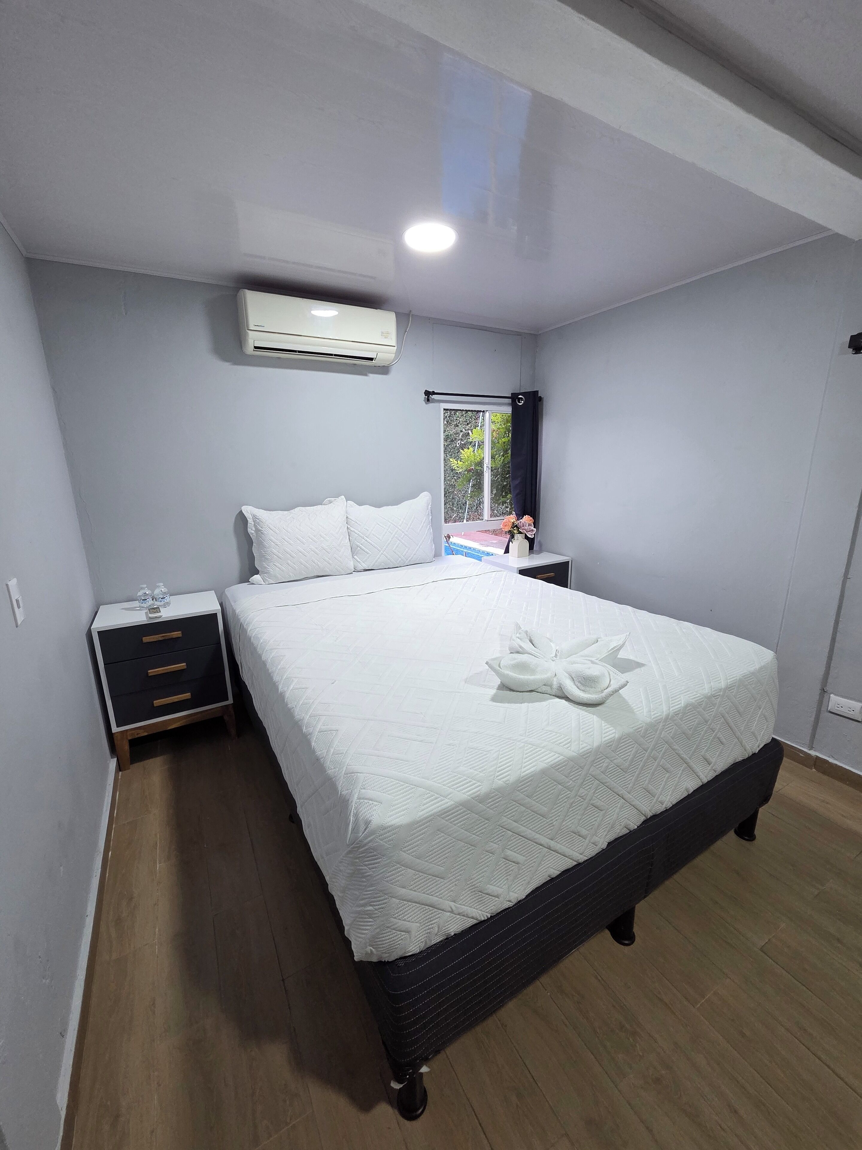 A bedroom with a bed, a nightstand, a window with a view, and an air conditioning unit.