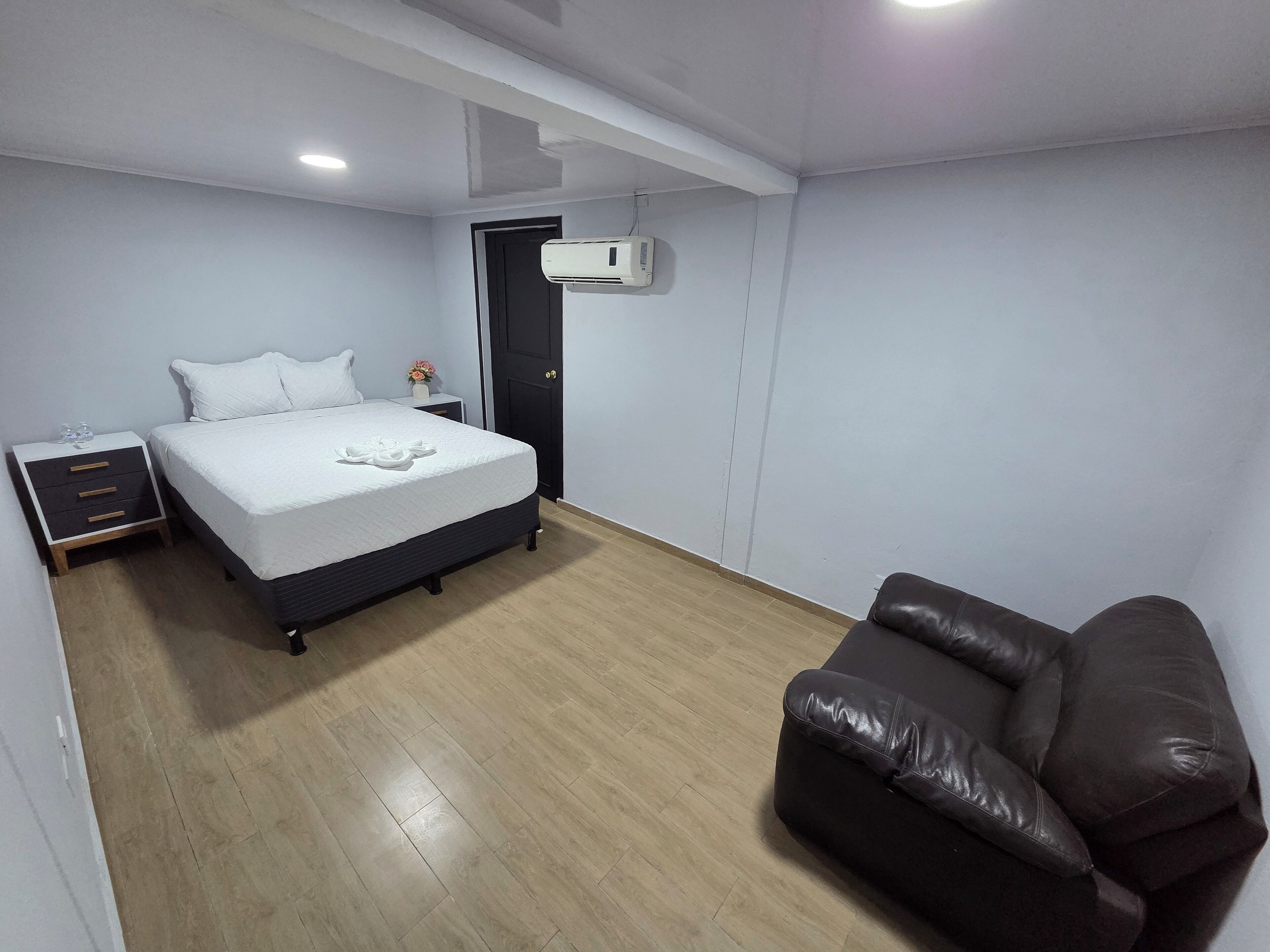 Une chambre à coucher comprenant un lit, une table de chevet, une commode, un canapé en cuir marron et un climatiseur fixé au mur.