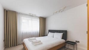 Una moderna camera d'albergo con un letto in legno, lenzuola bianche, un piccolo comodino e una finestra con tende trasparenti.