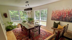 A dining area with a wooden table, chairs, and a red patterned rug. There are large windows with a view of greenery outside. - Etowah Valley Ranch - Peaceful, private, and serene!  CLose to everything! (Dahlonega)
