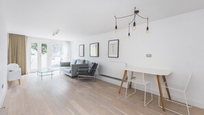 A modern, well-lit room with a large window, a grey sofa, a white table, and a hanging light fixture.