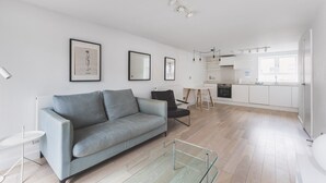 A modern apartment with a grey sofa, a glass coffee table, a kitchen with white cabinets, and framed artwork on the walls.
