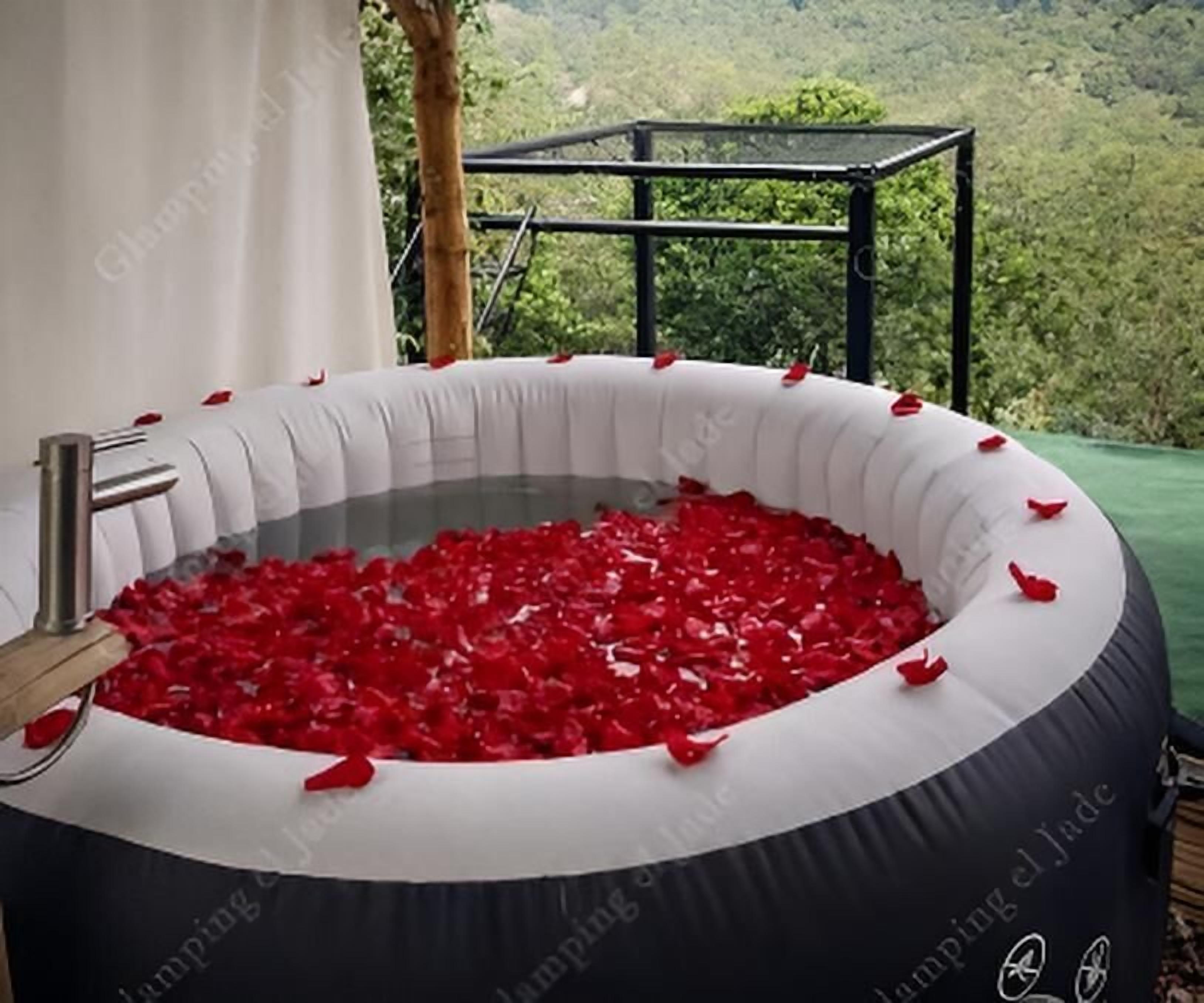 A hot tub filled with red rose petals, surrounded by a white and black inflatable structure, with a view of greenery in the background.