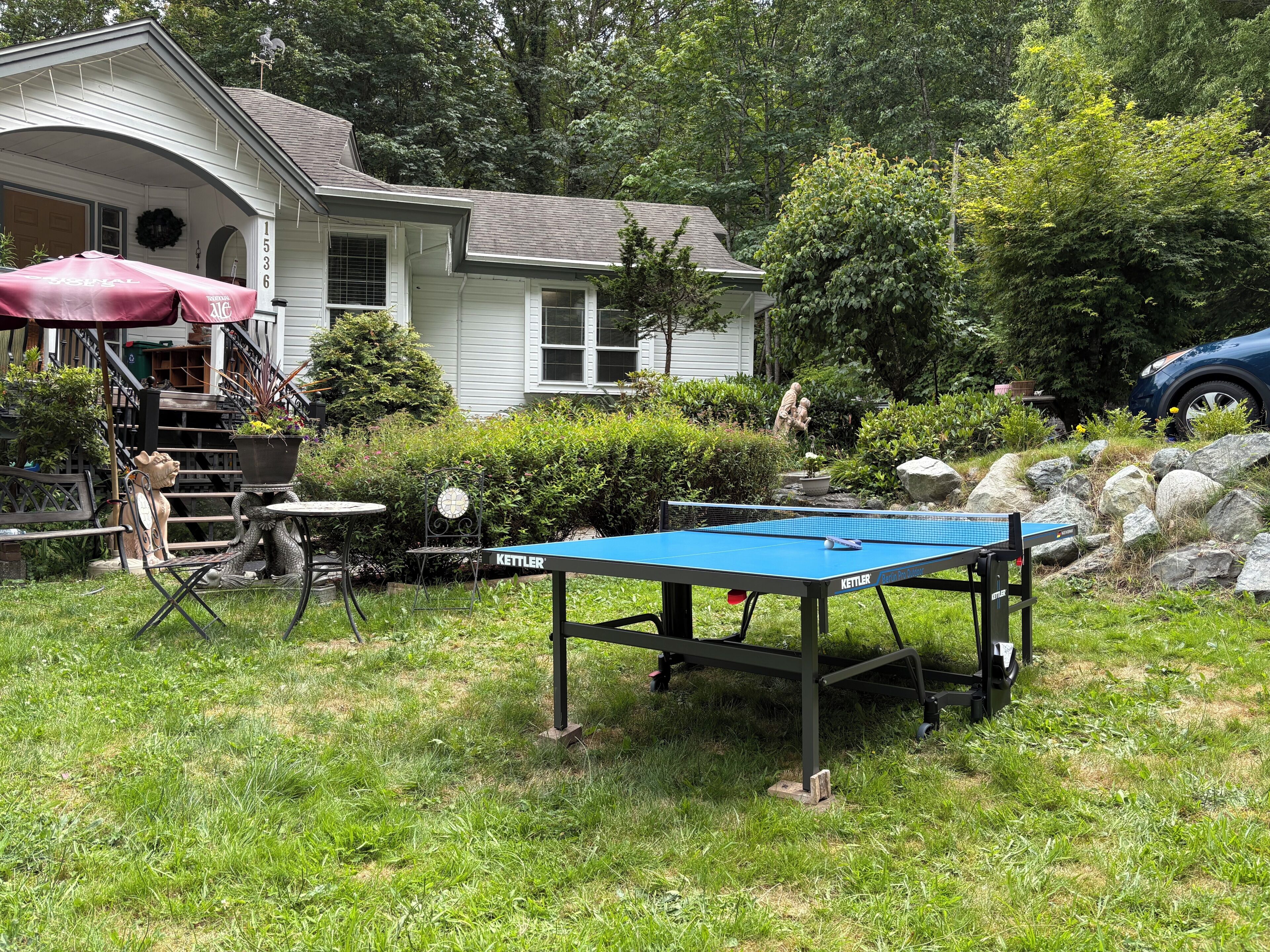 A residential setting with a blue ping pong table, outdoor seating, and a house with a porch.