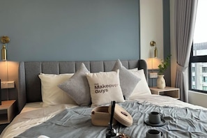 Une chambre d’hôtel avec un lit, des oreillers, une table de chevet, une lampe, un vase avec des fleurs et une fenêtre avec des rideaux.