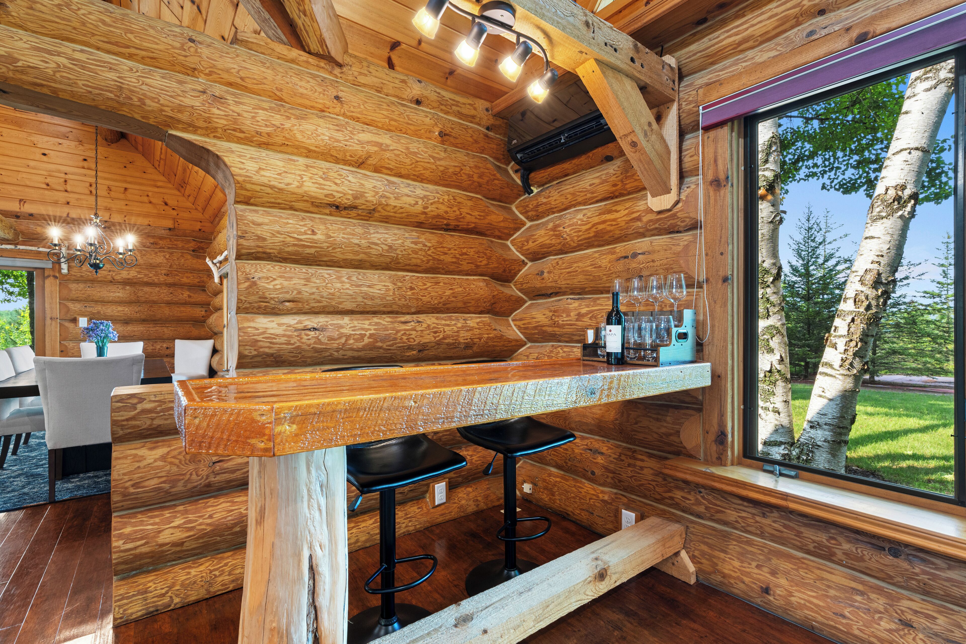 Interior de una cabaña de troncos con una barra de madera, dos taburetes y vistas a árboles de abedul.