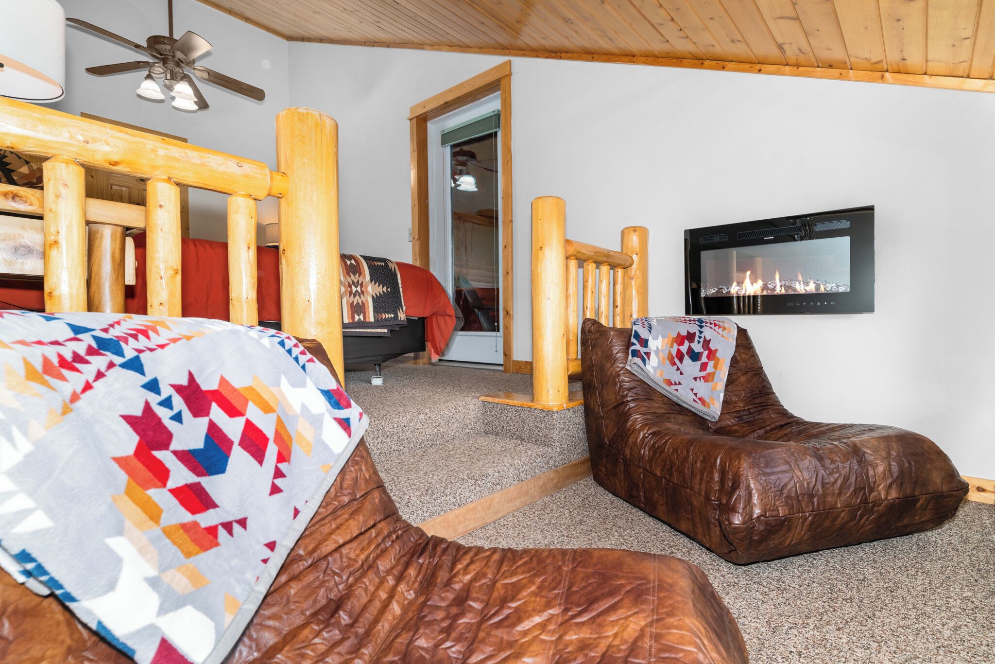 A cozy room with a wooden ceiling, a bed with a colorful quilt, a leather chair, and a fireplace with a TV mounted above it.