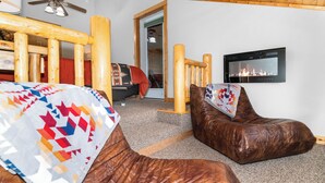A cozy room with a wooden ceiling, a bed with a colorful quilt, a leather chair, and a fireplace with a TV mounted above it.