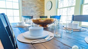 A dining table set with white plates, bowls, cutlery, and glasses, with a rustic brick wall and large windows in the background. - Quarter 54 (Palmer)