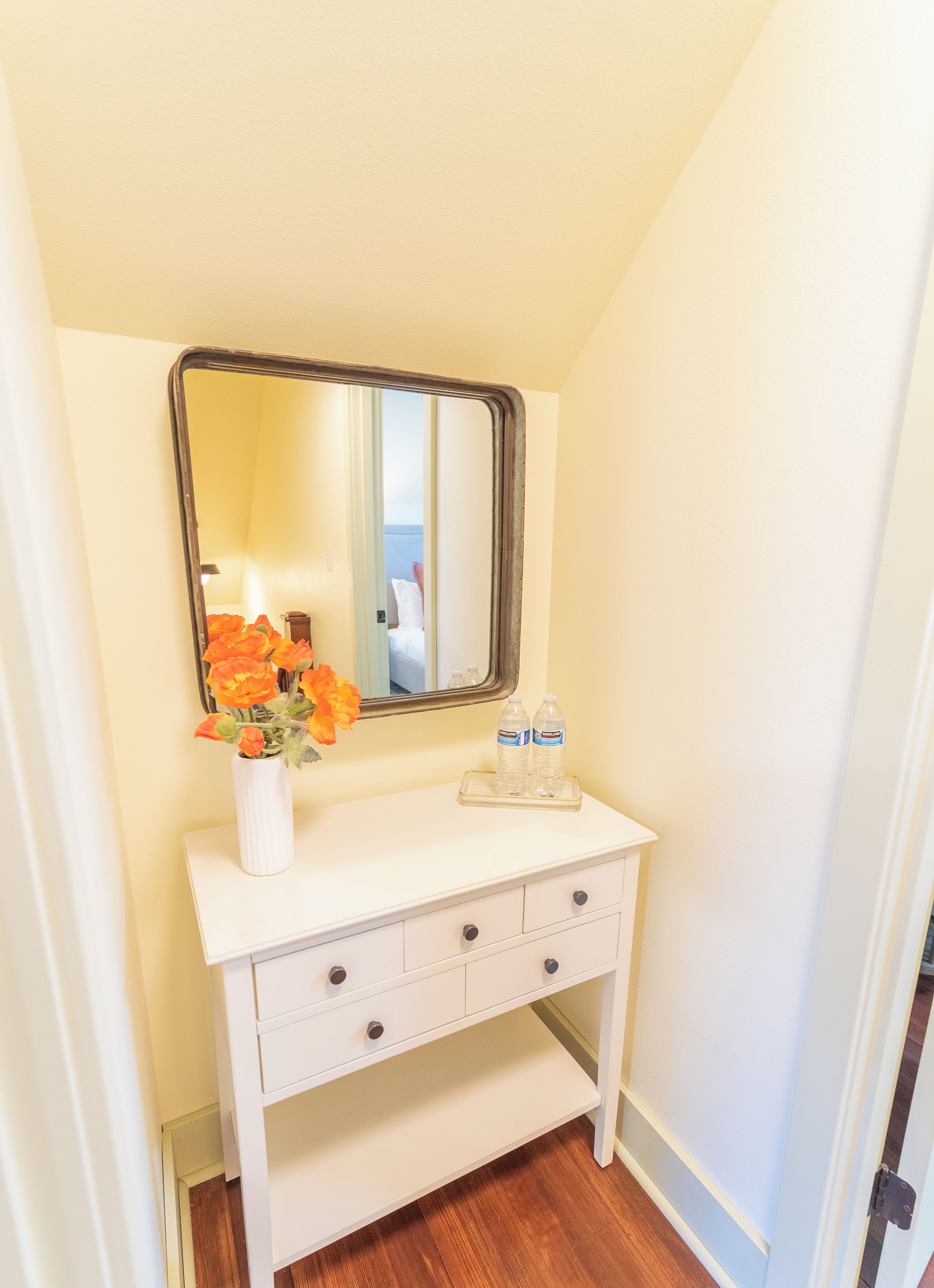 A bathroom with a white vanity, a mirror, and a vase with flowers. There are two bottles of water on the vanity.