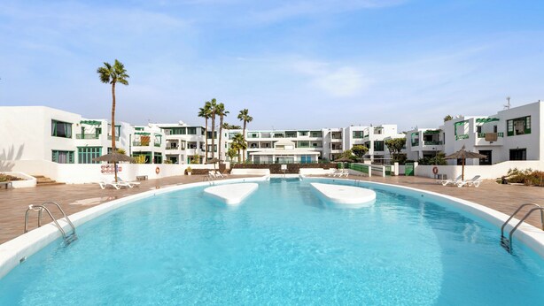 A swimming pool with lounge chairs and palm trees, surrounded by white buildings with balconies.