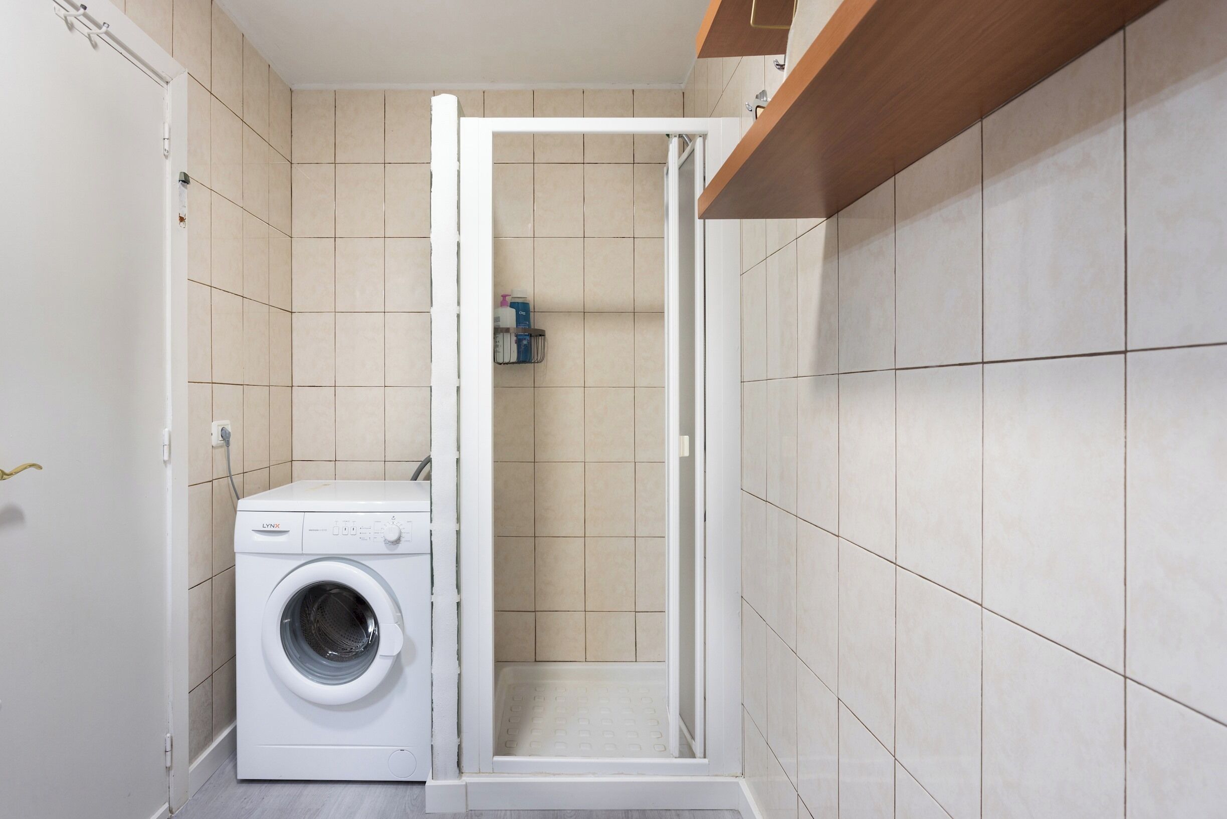 Une petite buanderie équipée d’un lave-linge à chargement frontal, d’un espace douche et d’une étagère pour les articles de toilette.