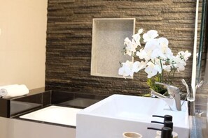 A modern bathroom with a stone wall, a large mirror, and white orchids. - VIlla Avi (Bali)