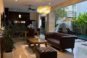 Un salon moderne avec un canapé, un pouf et une table à manger. Il y a également un espace piscine avec des chaises longues et un ventilateur de plafond.