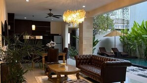 A modern living room with a leather sofa, a wooden coffee table, and a chandelier. There is a pool visible through the glass wall. - VIlla Avi (Bali)