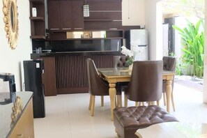 A modern kitchen with dark cabinetry, a central island, and a dining area with leather chairs and a glass table. - VIlla Avi (Bali)