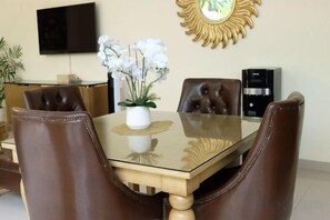 Un coin repas avec une table en verre, des fauteuils en cuir, un vase rempli de fleurs blanches, un miroir décoratif et un téléviseur fixé au mur.
