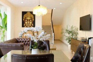 Un salon moderne avec une table à manger en verre, des fauteuils en cuir, une statue de Bouddha et un téléviseur fixé au mur.