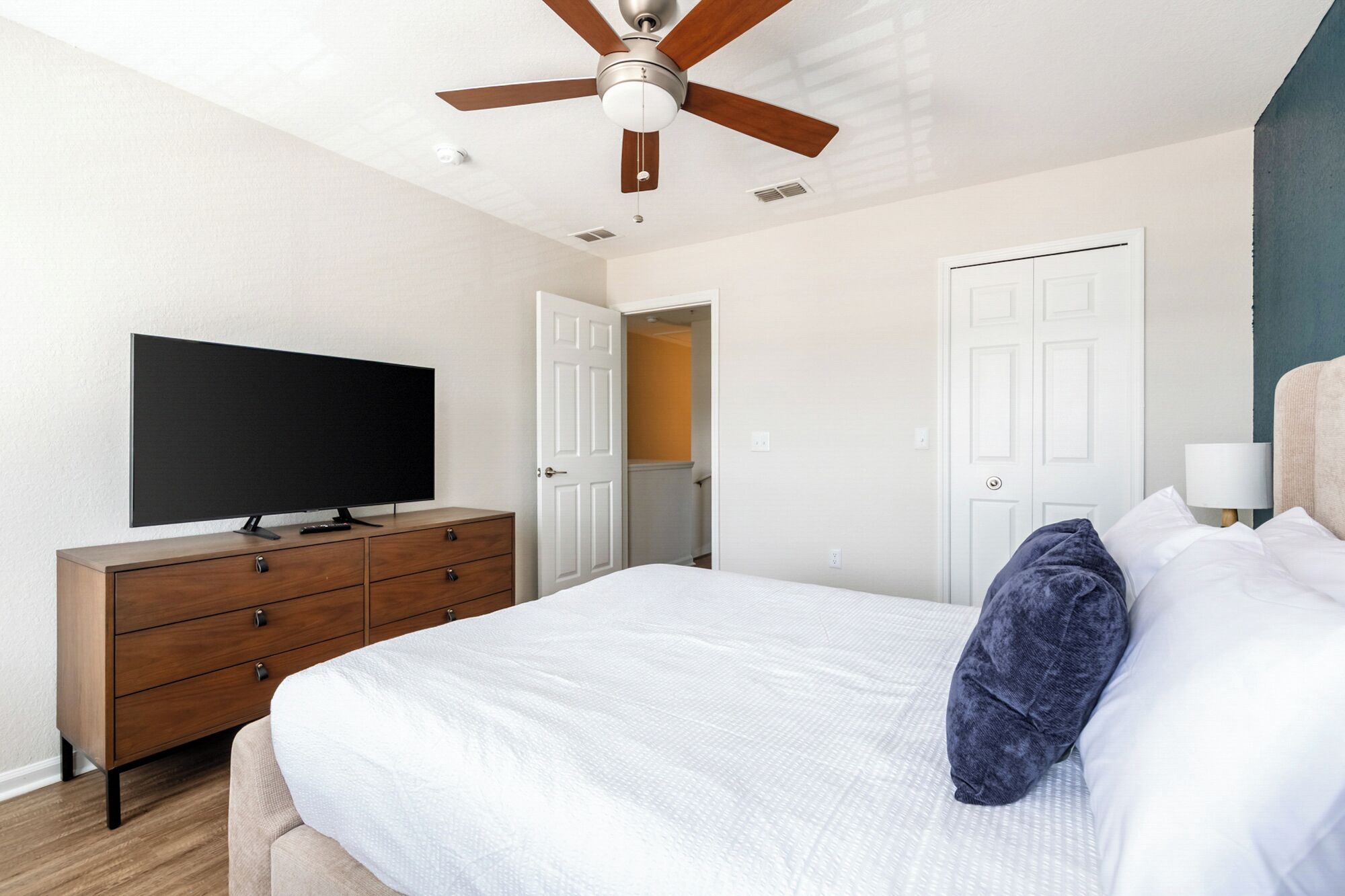 Une chambre à coucher comprenant un lit, une commode, un téléviseur, un ventilateur de plafond et une porte donnant sur une autre pièce.