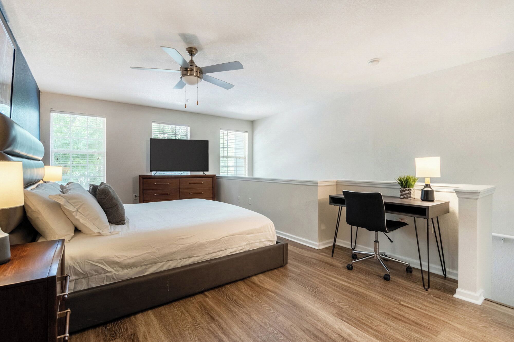 Ein Schlafzimmer mit Bett, Schreibtisch, Stuhl, Fernseher, Deckenventilator und Fenstern.