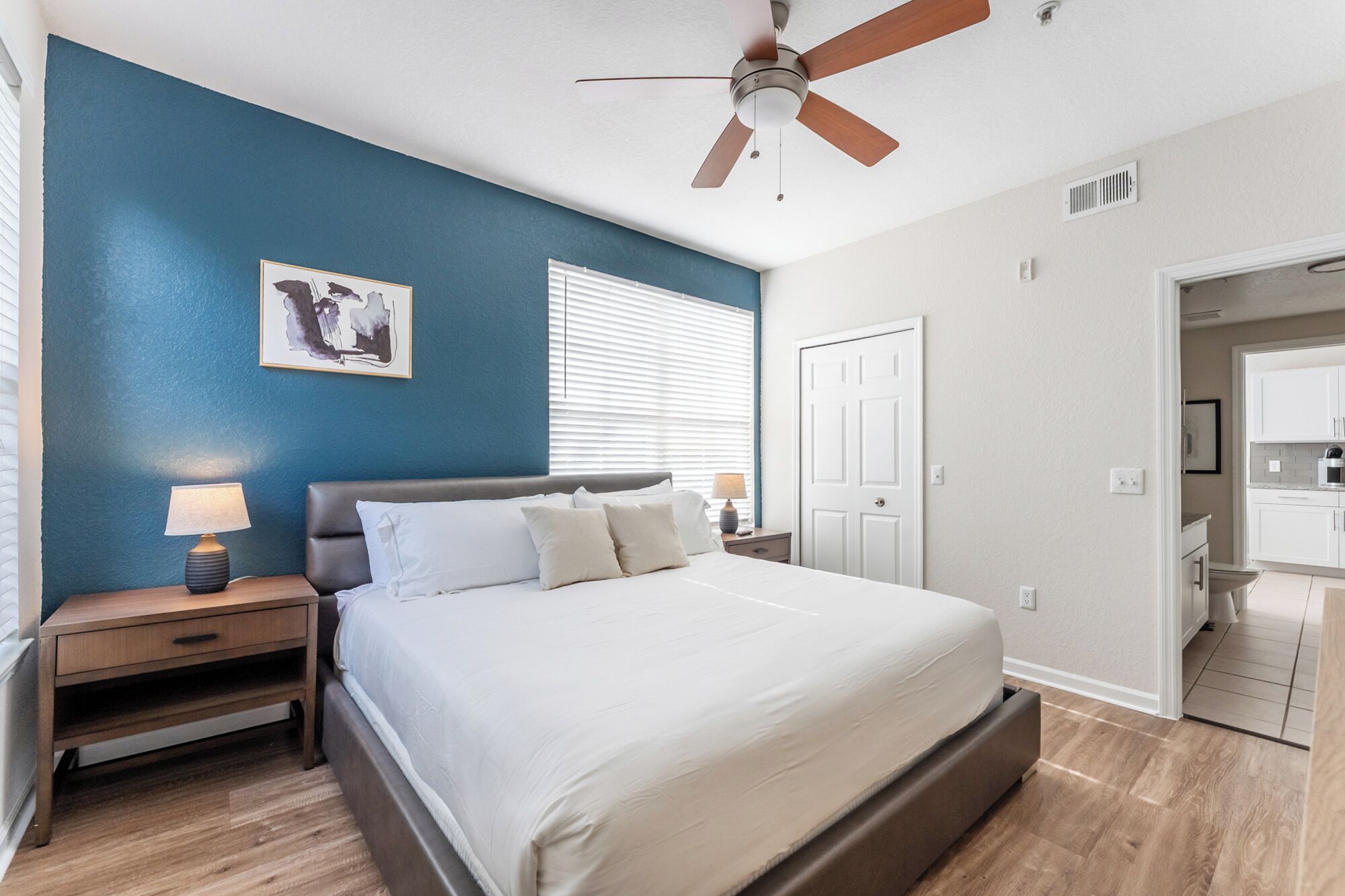 A bedroom with a bed, nightstands, a ceiling fan, a window with blinds, and a door leading to a bathroom.