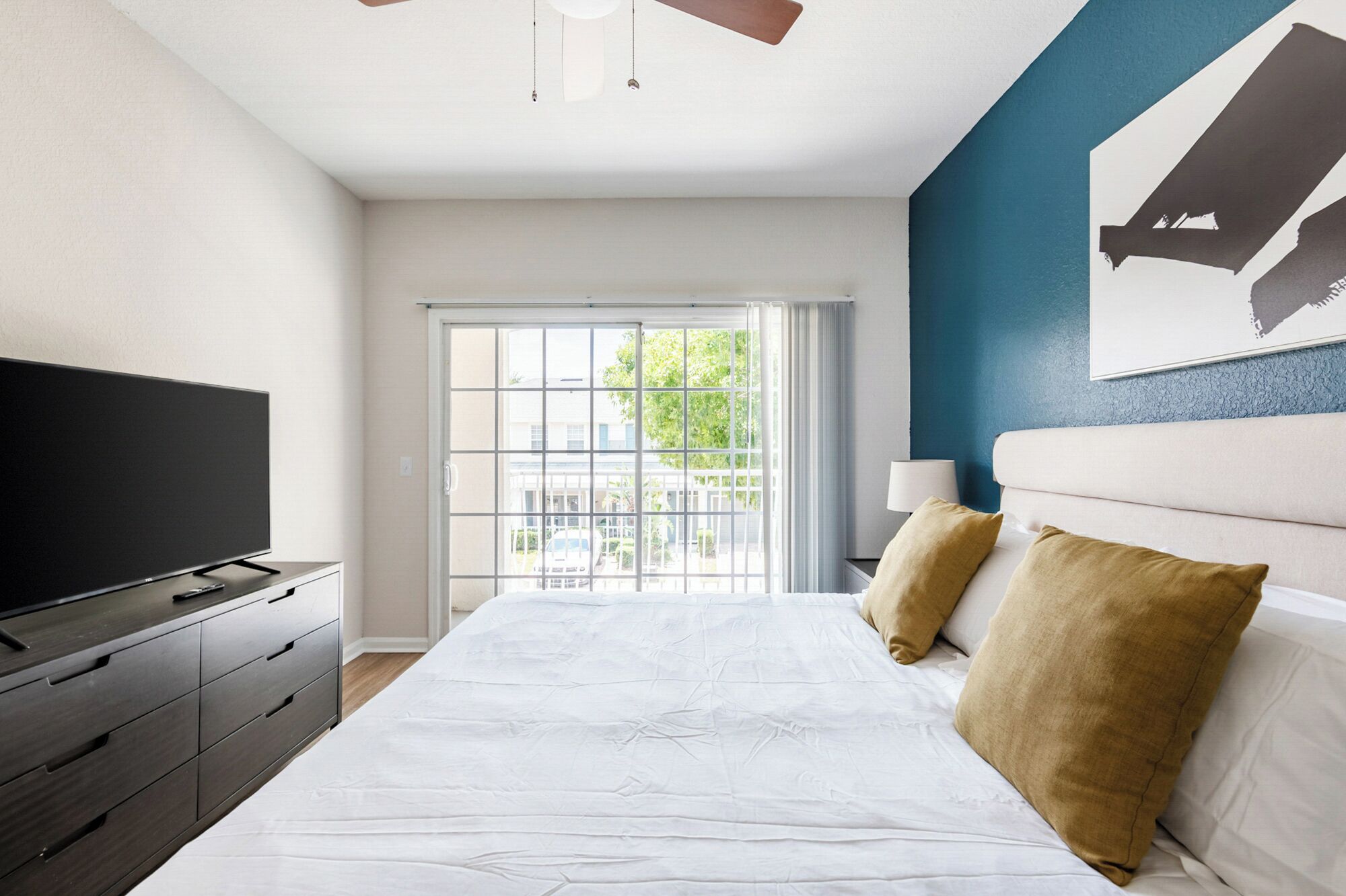 A modern bedroom with a large bed, a flat-screen TV, a dresser, and a view of the outdoors through a sliding glass door.