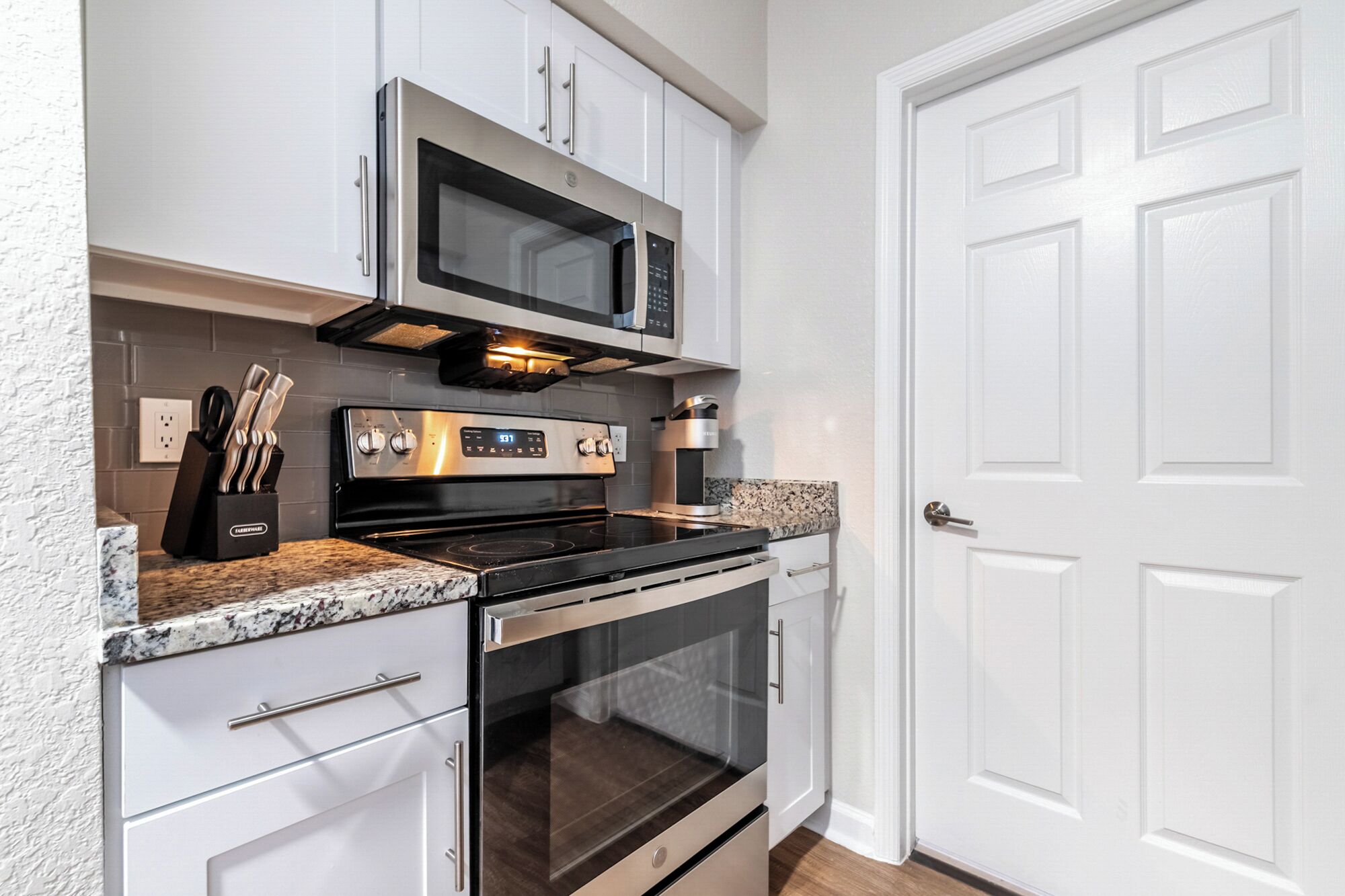 A kitchen with a stainless steel stove, microwave, and oven, white cabinets, granite countertops, and a white door.