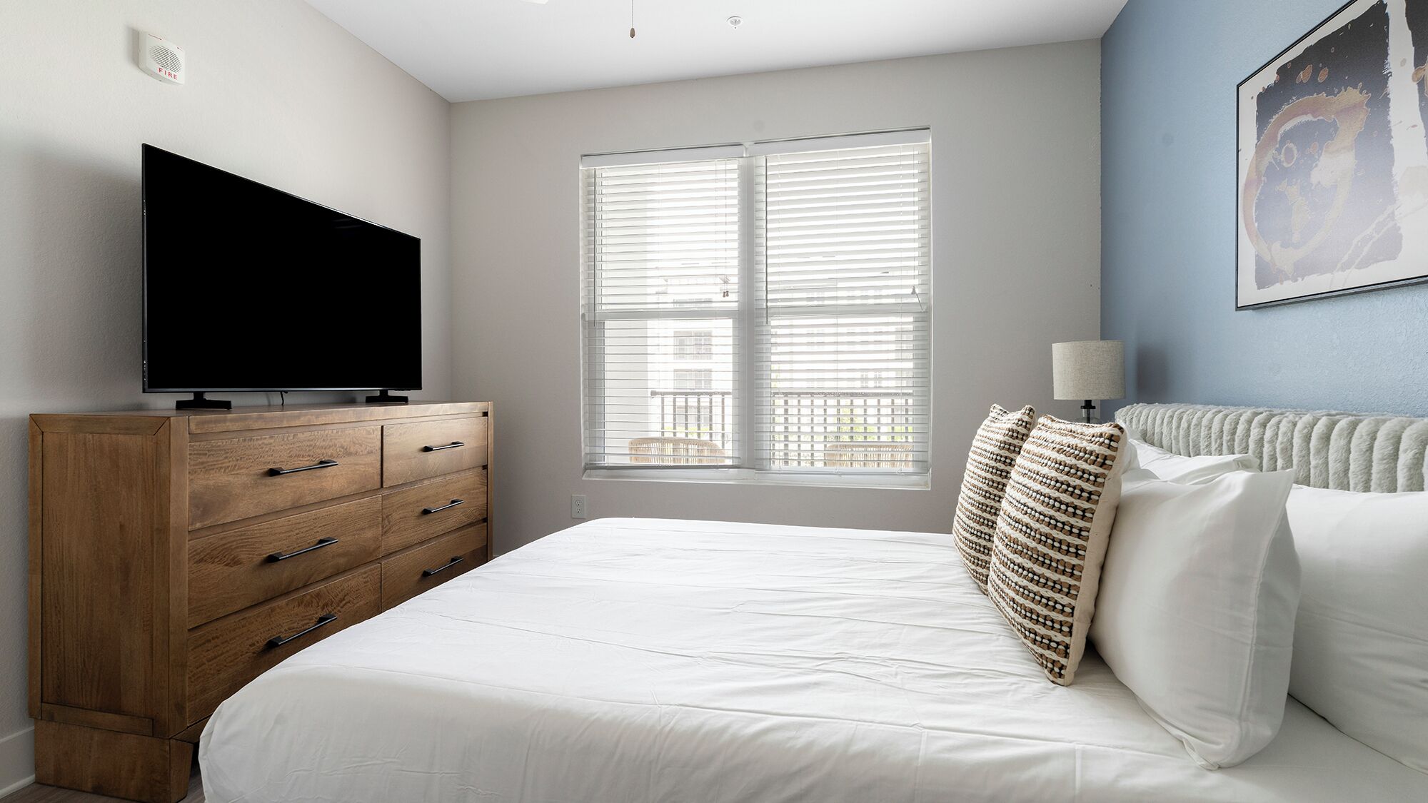 Un dormitorio con cama, cómoda de madera, televisor de pantalla plana, ventana con persianas y lámpara de pared.
