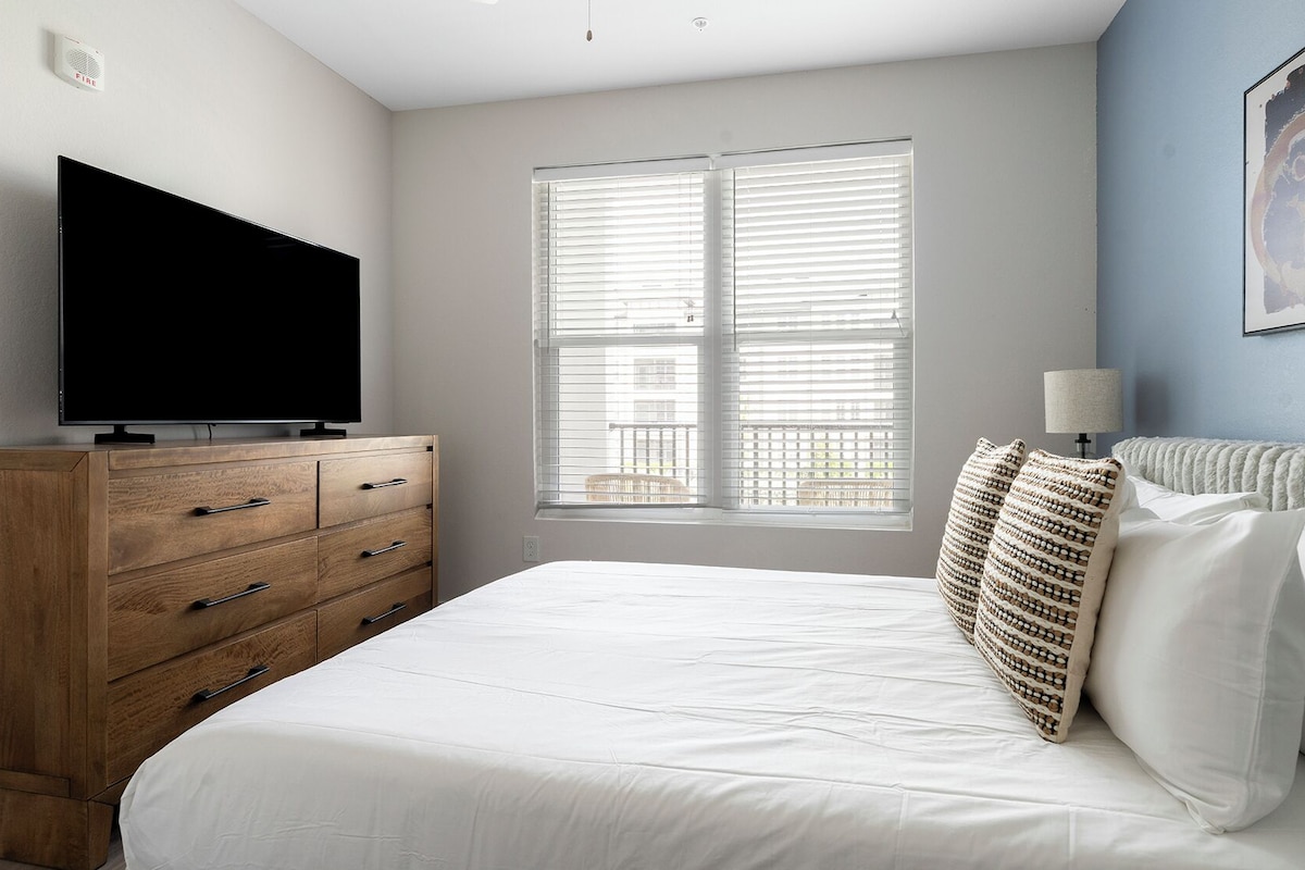 A bedroom with a bed, a wooden dresser, a flat-screen TV, a window with blinds, and a wall-mounted lamp.