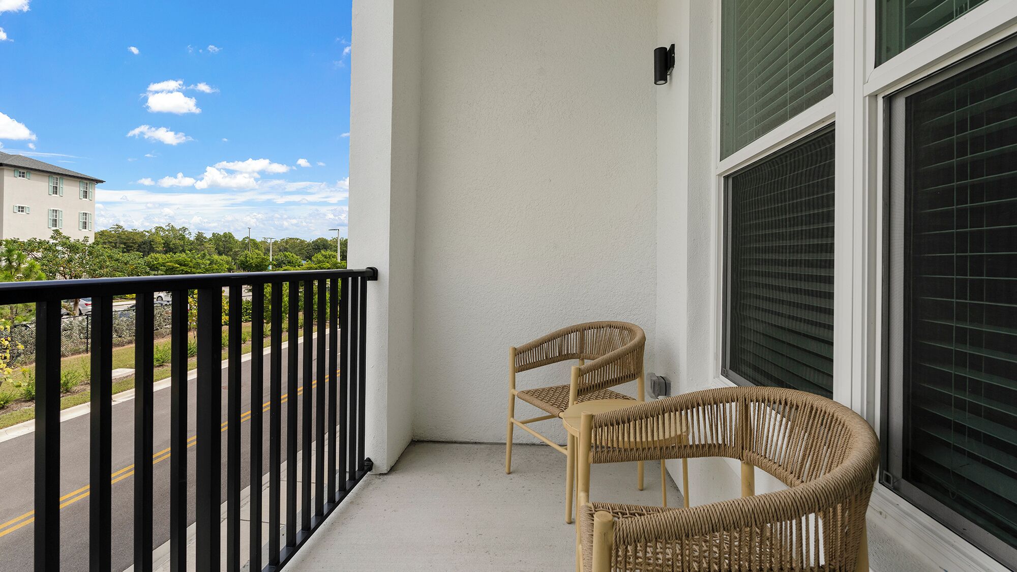 A balcony with two wicker chairs, a black railing, and a view of a road and greenery.