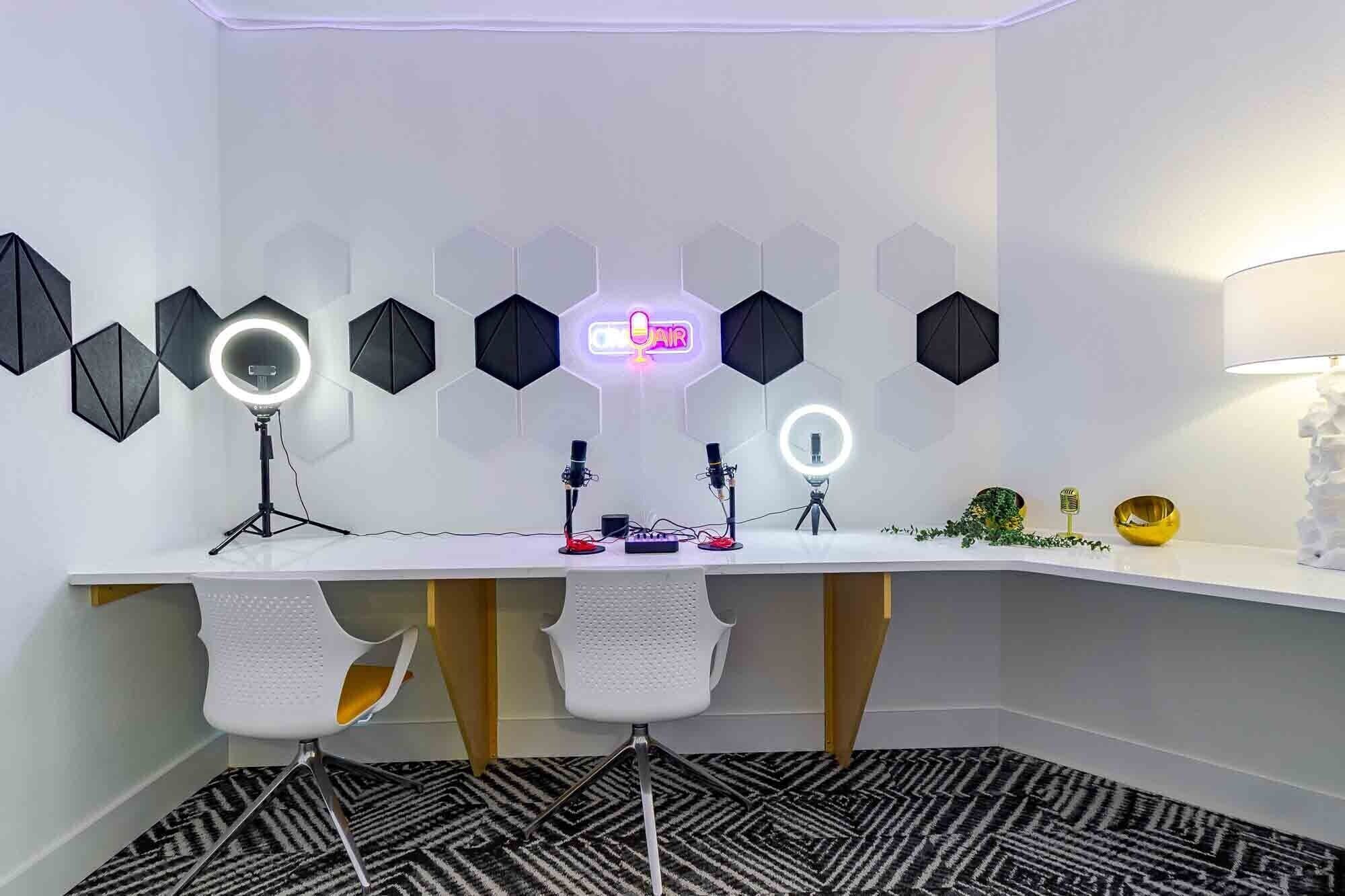A modern recording studio with a white desk, two white chairs, hexagonal acoustic panels, and two ring lights.
