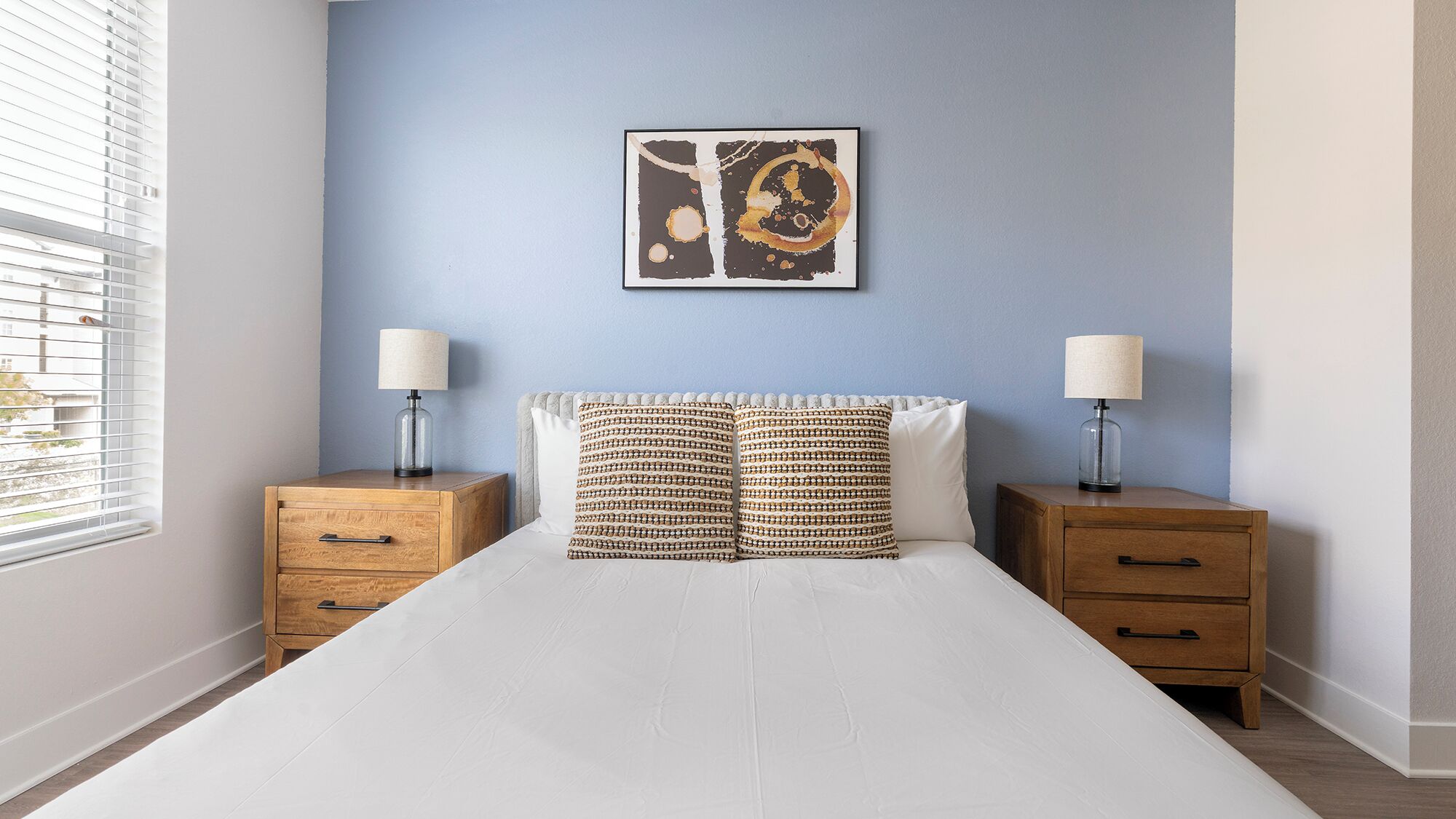 Una camera da letto con un letto, due comodini, una lampada su ciascuno e un quadro appeso al muro, oltre a una finestra con persiane.