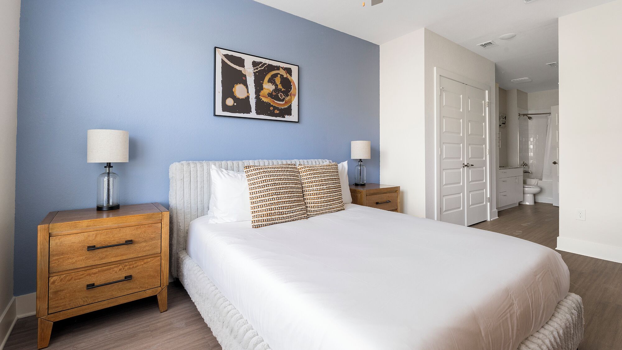 A bedroom with a bed, two bedside tables, a nightstand, a wall-mounted lamp, and a door leading to a bathroom.