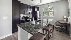 A modern kitchen with dark cabinets, a granite countertop, a stainless steel refrigerator, and a window with blinds. - Landing Apartments Charleston - Summerville (Summerville)