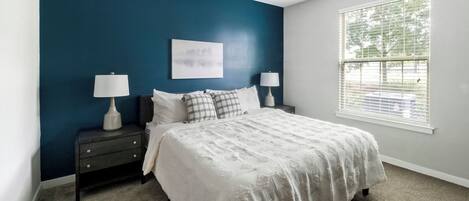 A bedroom with a bed, two nightstands, two lamps, a window with blinds, and a wall with a blue accent.
