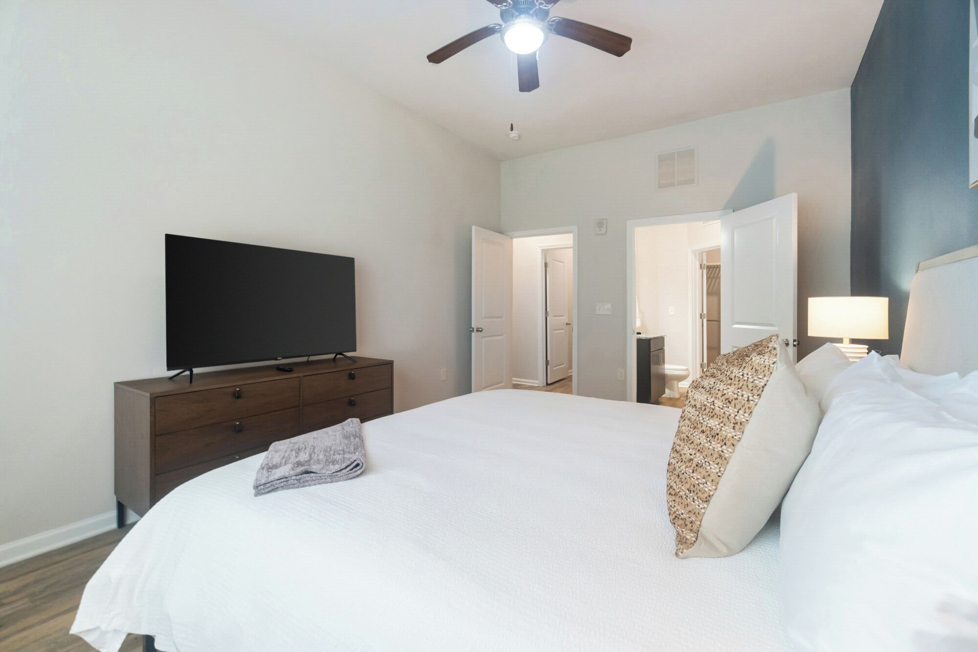 A hotel room with a large bed, a flat-screen TV on a wooden dresser, a ceiling fan, and a nightstand with a lamp.