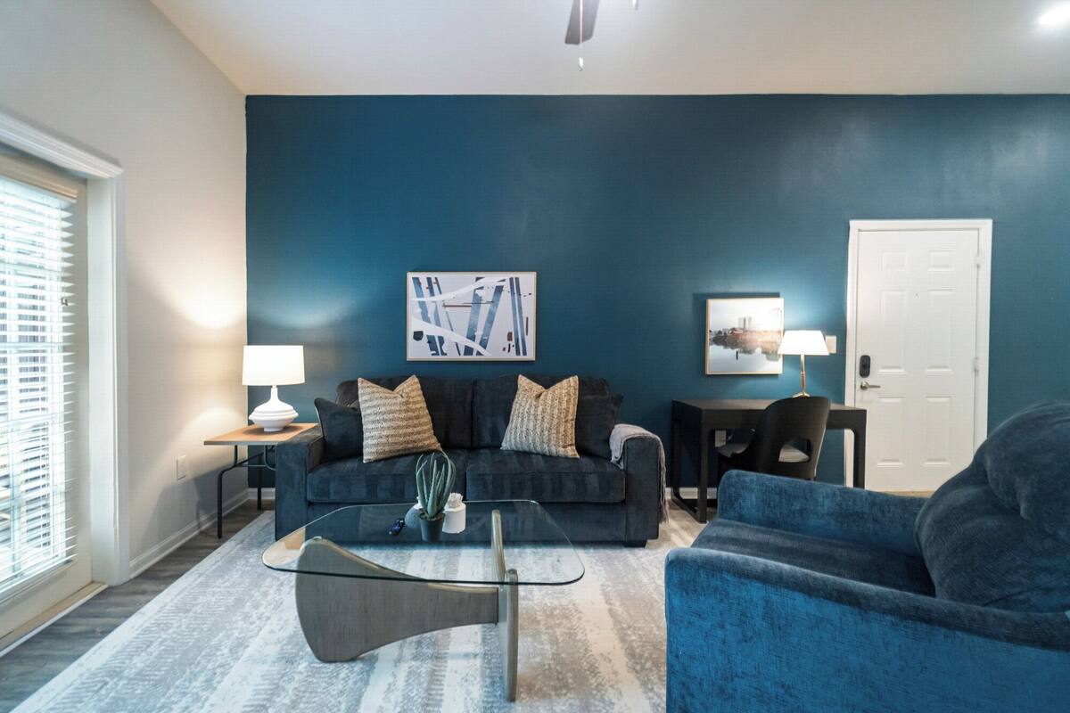 Un salon avec un mur bleu foncé, une table basse en verre, un canapé bleu foncé et deux tables d’appoint avec des lampes.