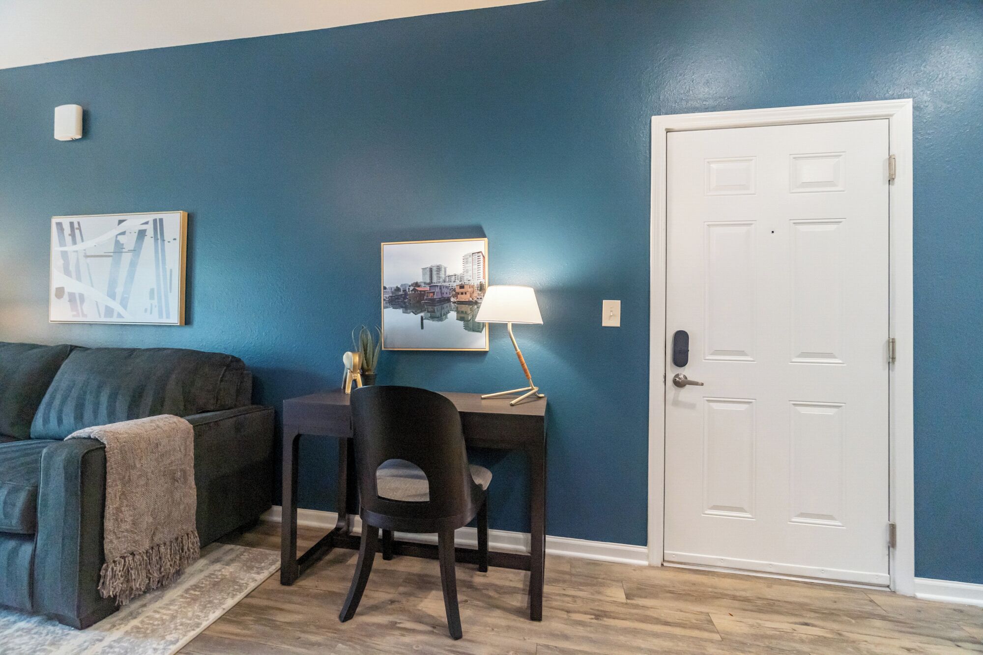 Une pièce avec un mur bleu, une porte blanche, un canapé de couleur foncée, une petite table avec une lampe et deux tableaux encadrés.